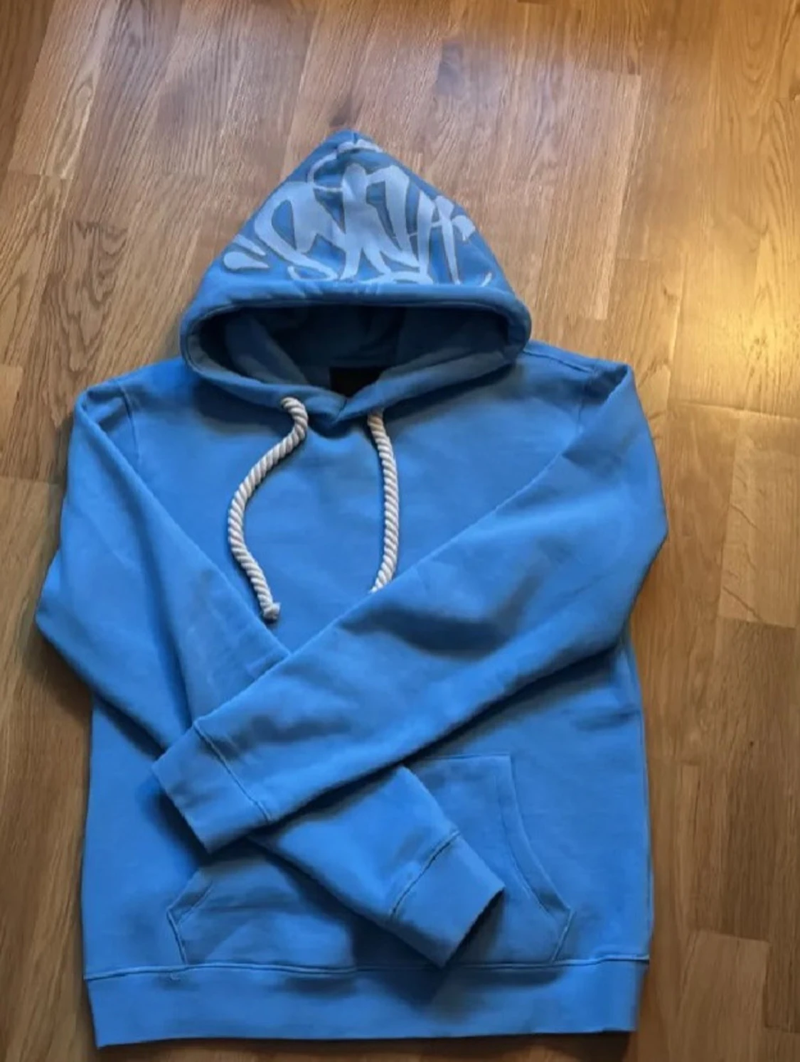 Ljusblå Streetwear Hoodie med Vit Logotyp• Trendig Ljusblå Hoodie med Graffiti-logga• Ljusblå Hoodie med Tryck på Huva• Modern Street Hoodie – Ljusblå• Casual Ljusblå Hoodie med Känguruficka