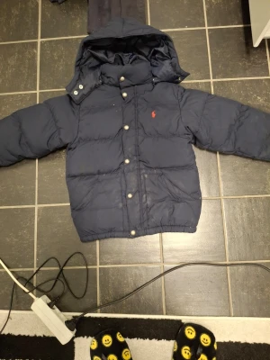 Mörkblå pufferjacka från Polo Ralph Lauren - Säljer en mörkblå pufferjacka från Polo Ralph Lauren med huva och silverfärgade knappar. Jackan har det klassiska röda Polo-logot broderat på bröstet och två stora fickor framtill. Perfekt för kalla dagar och riktigt snygg till streetwear.