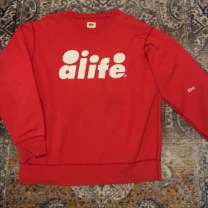 Röd sweatshirt från Alife - Röd sweatshirt från Alife med stor vit logga framtill och mindre logga på ärmen. Klassisk rund halsringning och långa ärmar. Perfekt för dig som gillar streetwear och vill sticka ut med en enkel men snygg design. Fint skick storlek Large