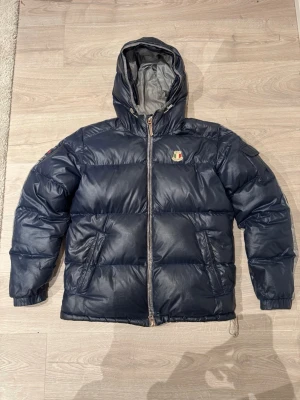 Mörkblå pufferjacka från Dolomite - Säljer en mörkblå pufferjacka från Dolomite med huva och broderade detaljer på bröstet, ryggen och ärmen. Jackan har dragkedja framtill, två snedställda fickor och elastiska muddar. Perfekt för kalla dagar och riktigt snygg streetstil.