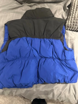 Blå och svart dunväst med tryckknappar - Säljer en blå och svart dunväst med hög krage och tryckknappar framtill. Västen har två sidofickor och elastiska sömmar som ger en puffig look. Perfekt för lager på lager under kyliga dagar.