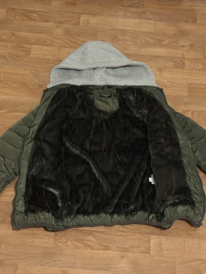 Grön pufferjacka med grå luva - Säljer en grön pufferjacka från Hello Boy med grå luva i sweatshirtmaterial. Jackan har svart fuskpälsfoder på insidan, dragkedja framtill och två sidofickor med dragkedja. Perfekt för kalla dagar och riktigt skön att bära.