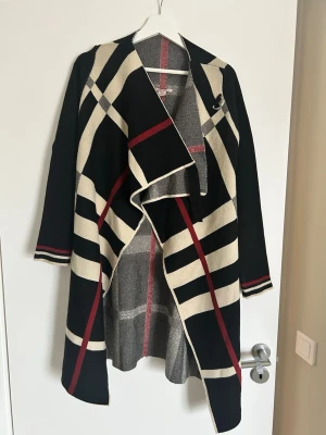 Rutig lång cardigan kofta från Anna Field - Säljer en lång cardigan kofta från Anna Field med svart, beige och rödrutigt mönster. Jackan har öppen framsida med snyggt fall, långa ärmar och dekorativa ränder vid ärmsluten. Perfekt för dig som gillar statement-plagg och vill ha något extra till höst och vinter.