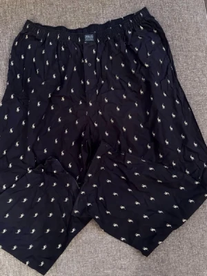 Svarta pyjamasbyxor från Polo Ralph Lauren - Säljer ett par svarta boxershorts från Polo Ralph Lauren med klassiskt mönster av små vita polospelare över hela tyget. De har resår i midjan och knappgylf. Perfekta för dig som gillar stilrena underkläder med en ikonisk touch.