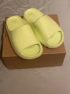 Adidas Yeezy Slide glow green - Yeezy Slides i ljusgrön färg. Storlek 38. Skorna är använda ett fåtal gånger och är i mycket fint skick, inga sprickor eller större slitage. Rena och väl omhändertagna. 100 % äkta. Säljs då de tyvärr är för små för mig och därför inte kommer till användning. Skickas snabbt. Perfekta till chil dagar!