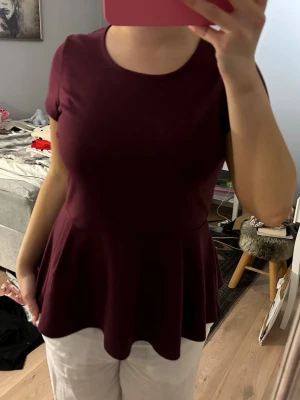 Vinröd peplum topp med kort ärm - Säljer en vinröd peplum topp med rund halsringning och korta ärmar. Toppen har en markerad midja och ett utsvängt nedre parti som ger en snygg siluett. Tillverkad i mjukt och stretchigt material, perfekt till jeans eller kjol.