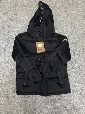 Svart pufferjacka från Burberry - Säljer en svart pufferjacka från Burberry med avtagbara ärmar så den kan användas som väst. Jackan har huva med klassiskt rutigt foder, två dragkedjefickor och Burberry-logga på ärmen. Perfekt för kalla dagar och riktigt snygg streetstil.