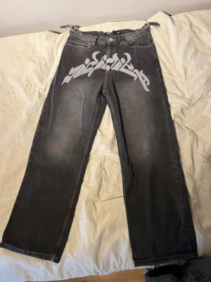Svart jeans från Vicinity med grått embroidery på framsidan - Säljer ett par svarta baggy jeans med stort grått embroidery över framsidan. Jeansen har vida ben och knytband i midjan för extra streetwear-vibe. Perfekta för dig som gillar unik och edgy stil. Köpta från Vicinity’s hemsida i 2023 och aldrig använda sen dess. Prislapp (se bild) och kvitto finns.