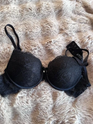 Svart spets-bh med rosett och pärla - Snygg svart bh med vadderade kupor och vacker spetsdetalj över hela ytan. Mellan kuporna sitter en liten rosett med en dekorativ pärla. Axelbanden är justerbara och bh:n har hakknäppning bak. Perfekt för dig som gillar en elegant och feminin stil.