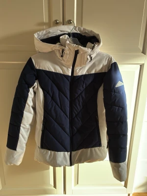 Skidjacka med huva från McKinley - Snygg skidjacka från McKinley i marinblått och vitt. Jackan har huva, dragkedja framtill och två sidofickor med dragkedja. Quiltad design med diagonala sömmar och logga på ärmen. Storlek 36. I fint skick och nytvättad. 