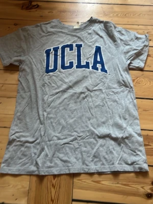 Grå UCLA t-shirt från H&M - Snygg grå t-shirt från H&M med stor blå UCLA-tryck på bröstet. Klassisk rund halsringning och korta ärmar. Perfekt för en avslappnad och sportig stil. Tillverkad i mjukt bomullsmaterial.