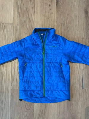 Blå lättviktsdunjacka barn från Norrøna - Snygg blå lättviktsdunjacka för barn från Norrøna med grön dragkedja och två fickor framtill. Jackan har hög krage och quiltad design, perfekt för kalla vinterdagar och som foder till en vindjacka. Lätt och smidig att bära. Passar för friluftsliv och skidåkning. 