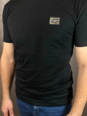 Svart t-shirt från Dolce & Gabbana - Stilren svart t-shirt från Dolce & Gabbana med en metallbricka på bröstet där loggan står. Klassisk passform och korta ärmar, perfekt till jeans eller under en jacka. Materialet känns mjukt och följsamt, troligen bomull.