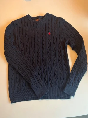 Mörkblå kabelstickad tröja från  Morris - Snygg mörkblå kabelstickad tröja från Morris med rund halsringning och diskret röd logga broderad på bröstet. Perfekt för en stilren och klassisk look. Tröjan är i storlek M men passar nog lika bra till S. Bara att höra av sig vid frågor. 
