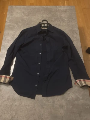 Mörkblå burberry skjorta - Stilren mörkblå skjorta med klassisk krage och långa ärmar. Insidan av manschetterna har snygga beige, röda och svarta rutiga detaljer som ger en unik touch. Skjortan har knappar framtill och är perfekt för dig som gillar en clean men ändå personlig stil.