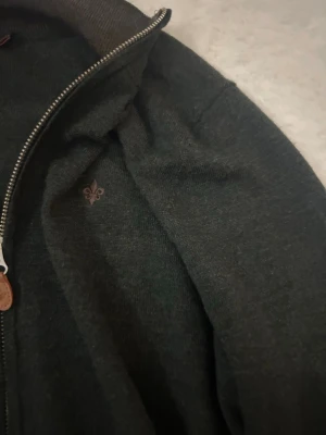 Mörkgrå ziptröja från Morris - Mörkgrön ziptröja från Morris med liten broderad logga på bröstet. Tröjan har hög krage, dragkedja med läderdetalj och är tillverkad i mjukt material. Perfekt för lager-på-lager och stilren look.