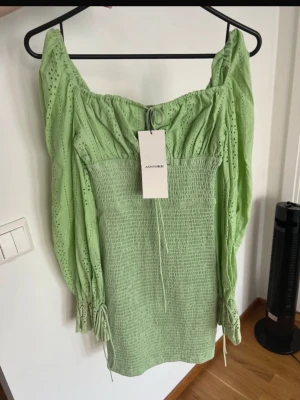 Grön kort klänning från Adoore - Snygg ljusgrön kort klänning från Adoore med smockad midja, broderade detaljer och långa puffärmar med knyt vid ärmslut. Klänningen har en söt offshoulder-ringning och volangdetaljer upptill. Perfekt för sommarens alla tillfällen!