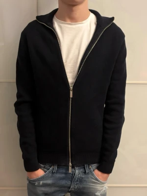 Mörkblå full-zip - Säljer en svart stickad kofta med hel dragkedja framtill. Tröjan har en enkel, clean look med ribbade muddar och krage. Perfekt till jeans och funkar året runt när du vill ha något stilrent och bekvämt.