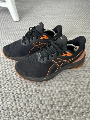 Svarta Asics sneakers med orange detaljer - Snygga svarta löparskor från Asics med orange detaljer och logga på sidorna. Skorna har, mesh-ovandel och GORE-TEX-material som gör dem vattentäta. Perfekta för löpning och träning utomhus. 