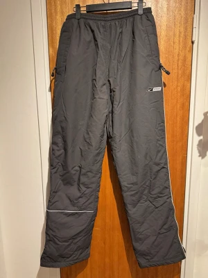 Grå täckbyxor från Nike - Täckbyxor i mörkgrått från Nike med resår i midjan och dragkedjor vid fickorna. Byxorna har vita detaljer längs benen och reflexband vid knäna. Perfekta för kalla dagar och outdoor-aktiviteter.