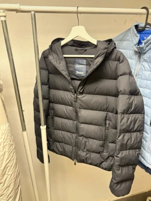 Svart pufferjacka från Herno - Snygg svart pufferjacka från Herno med huva och dragkedja framtill. Jackan har två praktiska fickor med dragkedja och quiltad design. Perfekt för kalla dagar och ger en stilren look. Materialet är lätt och isolerande.