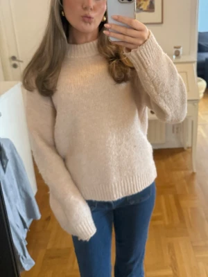 Rosa stickad tröja med rund hals - Rosa beige stickad tröja med rund hals och ribbade muddar vid ärmslut och nederkant. Tröjan har en avslappnad passform och långa ärmar, perfekt för lager på lager under kyliga dagar. Passar till både jeans och kjol.