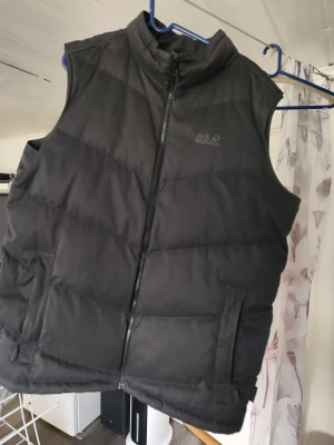 Svart dunväst från Jack Wolfskin - Stilren svart dunväst från Jack Wolfskin med hög krage och dragkedja framtill. Västen har två praktiska fickor med dragkedja och quiltad design som ger en sportig look. Perfekt för lager-på-lager och outdoor-aktiviteter.