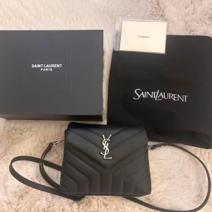 Svart Saint Laurent handväska - Säljer en svart handväska från Saint Laurent i quiltat läder med silverfärgad YSL-logga framtill. Väskan har justerbar axelrem och stängs med lock. Kommer med originalbox, dustbag och äkthetskort. Perfekt för dig som gillar lyxiga accessoarer.