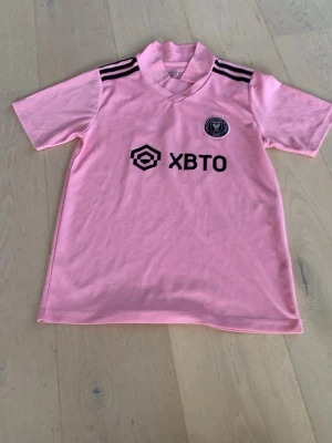 Inter Miami Messi matchtröja rosa - Rosa Inter Miami matchtröja från Adidas med svarta detaljer, klubbmärke och sponsortryck på bröstet. Messi och nummer 10 tryckt på ryggen. Korta ärmar och sportig passform, perfekt för fotbollsträning eller att bära som supporter. Materialet är lätt och andas.