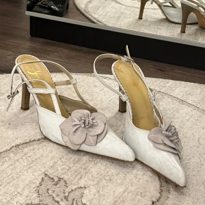 Vintage klackskor med blommig detalj - Säljer ett par eleganta vita klackskor med spetsig tå och en vacker blommig detalj på framsidan. Skorna har en smal rem runt ankeln. Perfekta för festliga tillfällen. Dessa är vintage.