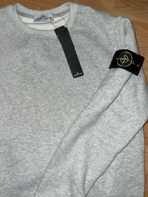 Grå sweatshirt från Stone Island - Snygg ljusgrå sweatshirt från Stone Island med klassisk rund halsringning och ikonisk svart logotyp-patch på ärmen. Tröjan har ribbade muddar vid ärmslut och nederkant. Perfekt för en clean och stilren look.