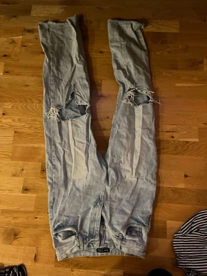 Ljusblå slitna jeans med hål - Säljer ett par ljusblå jeans med stora slitningar och hål vid knäna. Jeansen har raka ben och råa, avklippta benslut för en trendig och avslappnad look. Perfekta för dig som gillar streetstyle och en edgy vibe.