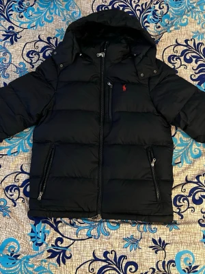 Svart jacka från Ralph Lauren - Svart dunjacka från Ralph Lauren med huva och dragkedja framtill. Jackan har två snedställda fickor med dragkedja och en liten röd logga på bröstet. Perfekt för kalla vinterdagar och riktigt snygg streetstil.