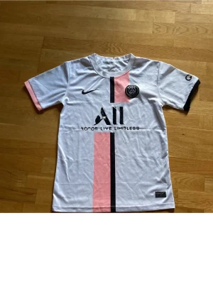 PSG matchtröja vit/rosa Nike - Snygg PSG matchtröja i vitt med rosa och svart vertikal rand. Tröjan har rund hals, korta ärmar och PSG-logga på bröstet. Tillverkad i lätt och ventilerande material, perfekt för fotbollsträning eller streetstyle.