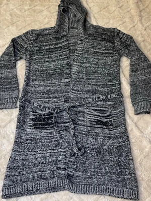 Gråmelerad stickad kofta med huva - Lång, gråmelerad stickad kofta med huva och knytskärp i midjan. Koftan har två framfickor och ribbstickade muddar vid ärmslut och nederkant. Perfekt för lager på lager och chill dagar.