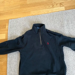 Svart half-zip tröja från Polo Ralph Lauren - Svart långärmad tröja från Polo Ralph Lauren med half-zip och klassisk röd broderad logga på bröstet. Tröjan har hög krage och är tillverkad i mjukt material som känns skönt mot huden. Perfekt för en stilren och avslappnad look.