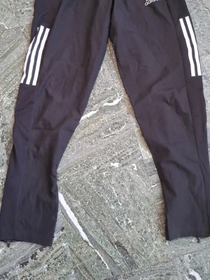 Svarta träningsbyxor Adidas S - Säljer ett par svarta träningsbyxor från Adidas med de klassiska vita ränderna längs benen. Byxorna är tillverkade i Primegreen-material och har elastisk midja. Perfekta för löpning eller gymmet.