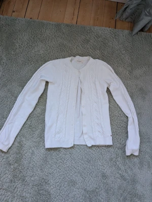 Vit kabelstickad kofta - Kabelstickad vit kofta med rund halsringning och knappar framtill. Långa ärmar och ribbade muddar vid ärmslut och nederkant. Klassisk och enkel design som passar till det mesta. Materialet känns mjukt och bekvämt.