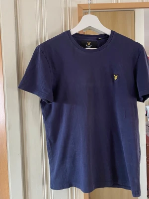 Mörkblå t-shirt från Lyle & Scott - Snygg mörkblå t-shirt från Lyle & Scott med klassisk rund hals och gul logga broderad på bröstet. T-shirten är i mjuk bomull och har en enkel, stilren design som passar till det mesta. STL S HERR