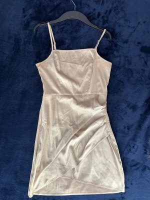 Champagnefärgad satin klänning - Snygg kort klänning i champagnefärgad satin med smala axelband och figurnära passform. Klänningen har omlottdetalj framtill och dragkedja i sidan för enkel påklädning. Perfekt för dig som vill ha en stilren och elegant look.
