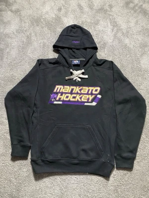 Svart Mankato Hockey hoodie - Snygg svart hoodie från J AMERICA med stort broderat Mankato Hockey-tryck i gult och lila på bröstet. Klassisk huva med vita snören och känguruficka framtill. Perfekt för dig som gillar hockey och vill ha en sportig look. Storlek S. Bra skick. Skriv om du har några frågor! Priset går att diskutera!