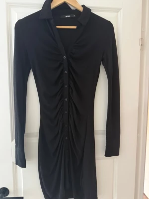 Svart skjortklänning med knappar - Svart långärmad skjortklänning från Bik Bok med knappar hela vägen framtill och snygg rynkdetalj längs mitten. Klänningen har klassisk krage och är figurnära i modellen. från BikBok, använd 1 gång