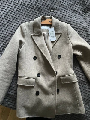 Beige kavaj från Donna, dubbelknäppt - Stilren beige kavaj från Donna med dubbelknäppning och svarta knappar. Klassisk krage, två fickor framtill och mjukt fodrad insida. Perfekt för en trendig och snygg look. Materialet är ullmix som ger en exklusiv känsla.