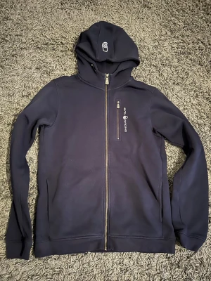 Marinblå hoodie från Sail Racing - Snygg marinblå hoodie från Sail Racing med dragkedja hela vägen, bröstficka med zip och logga på både bröstet och huvan. Perfekt för dig som gillar sportig och stilren look. Mjuk bomullskänsla och justerbar huva.