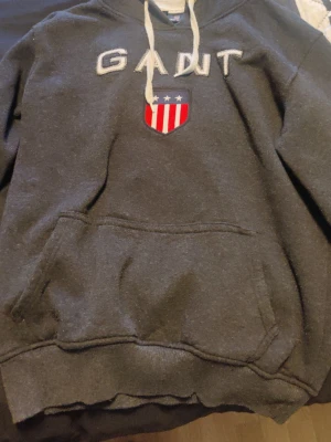 Mörkgrå hoodie från GANT stl L/XL - Mörkgrå hoodie från GANT med broderad logga och amerikanskinspirerat sköldmärke på bröstet. Klassisk känguruficka framtill och vita dragsnören i huvan. Perfekt för en avslappnad och sportig stil. Oanvänt skick 