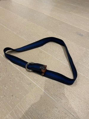 Randigt blått bälte Polo Ralph Lauren - Snyggt randigt bälte från Polo Ralph Lauren i marinblå och mörkblå nyans. Bältet har dubbla silverfärgade ringar som spänne och detaljer i brunt läder. Perfekt accessoar för att lyfta din outfit. Org pris: 800kr