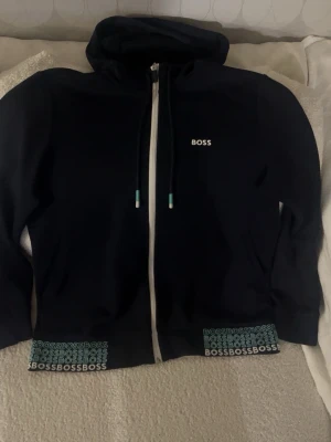 Svart hoodie med dragkedja från BOSS - Snygg svart hoodie från BOSS med vit dragkedja och huva. Diskret BOSS-logga på bröstet och upprepade BOSS-tryck längs nederkanten. Justerbara snören i huvan med turkosa detaljer. Perfekt för en avslappnad och trendig look.