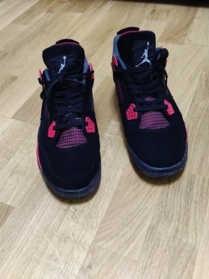 Svarta och röda Air Jordan 4 sneakers - Säljer ett par Air Jordan 4 sneakers i svart med röda och vita detaljer. Skorna har meshpaneler, klassiska snörningar och Jumpman-logga på plös och häl. Ovandelen är i mocka och textil, och insidan är fodrad i rött tyg. Perfekta för dig som gillar streetwear. Alla skador har jag tagit med på bilderna så kolla noga. Hör av dig vid frågor.