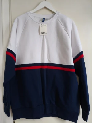 Vit och marinblå sweatshirt H&M - Snygg sweatshirt från H&M i vitt och marinblått med röda och mörkblå ränder över bröstet och ärmarna. Tröjan har rund halsringning och är tillverkad i mjuk bomullsmix. Perfekt för en sportig och avslappnad stil.