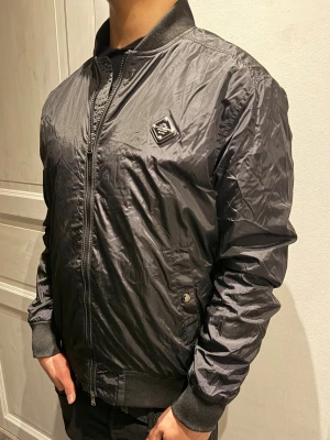 J.Lindeberg jacka  - Snygg svart bomberjacka från J.Lindeberg med glansigt nylontyg, ribbade muddar och krage. Jackan har dragkedja framtill, två fickor med knapp och en ikonisk Prada-logga på bröstet. Perfekt för dig som vill ha en stilren och trendig look.